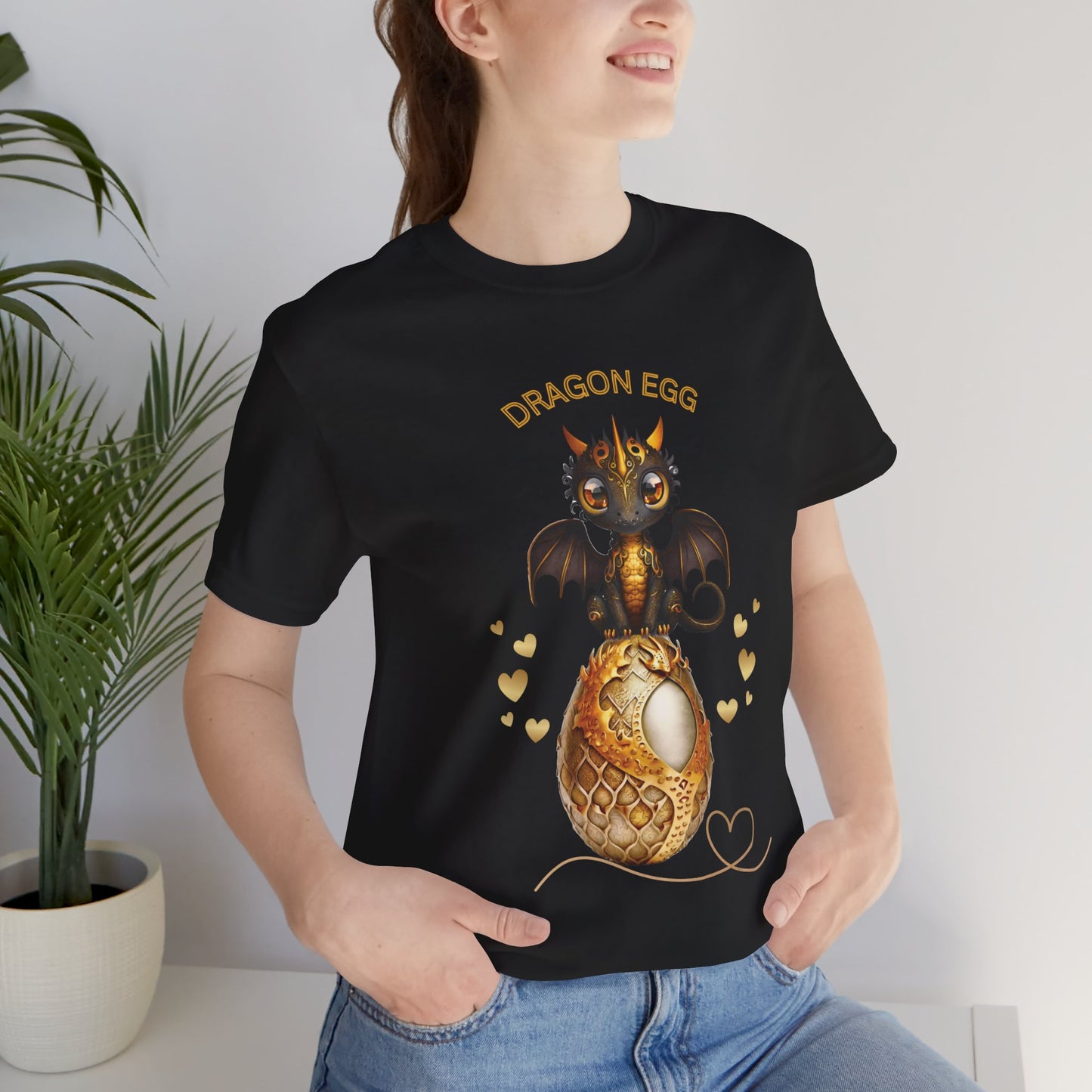A golden dragons egg with a brown & gold cute baby dragon with gold horns & wings out sits on top of her golden egg- hearts float up out of both sides of the egg - Text says Dragons Egg above the baby dragon and her golden dragon egg.