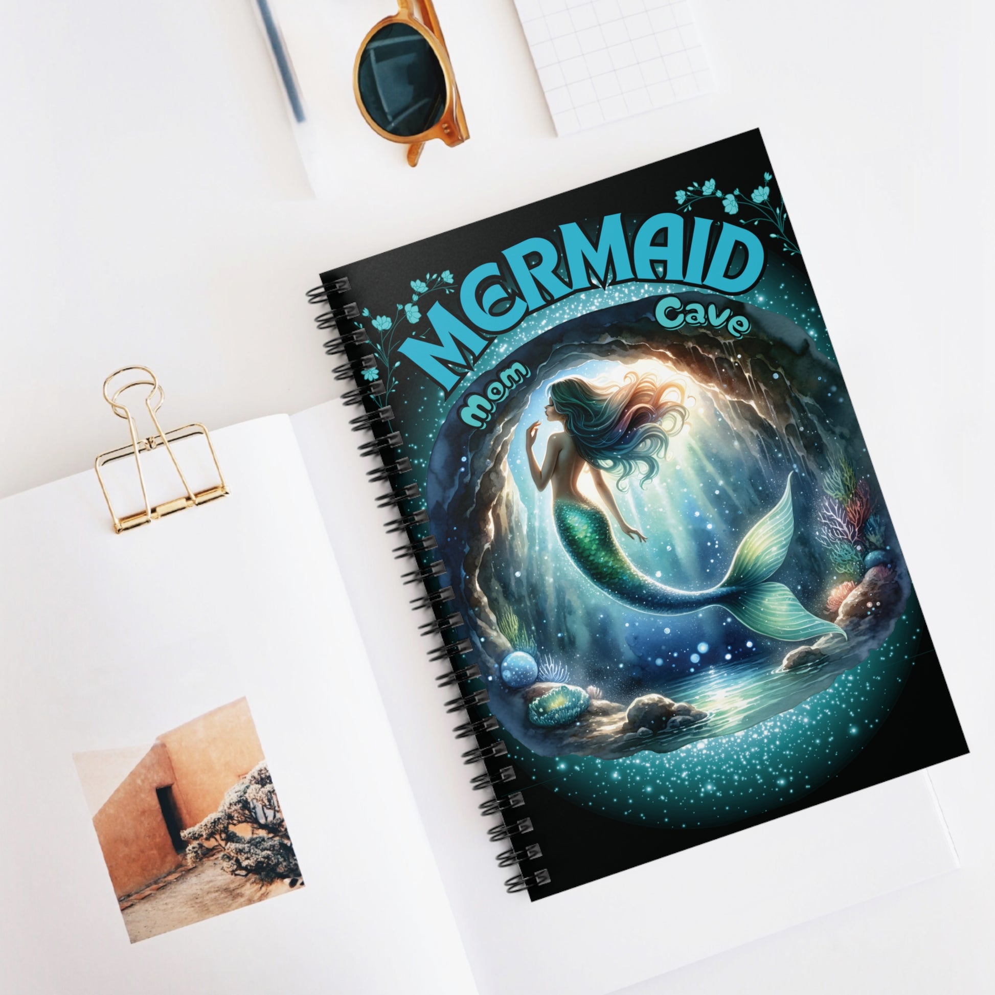 A gorgeous mermaid swims in her mermaid mom cave with her sea shells and plants around her - Mermaid Mom Cave is the text above her cave with blue flowers above the text - on the front cover of a lined spiral notebook