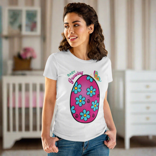 a pink egg with blue daisies with yellow centers and black squiggles inside the egg - Bbay Growing is the text - a blue and pink butterfly sits atop the egg on a short sleeve semi fitted t-shirt