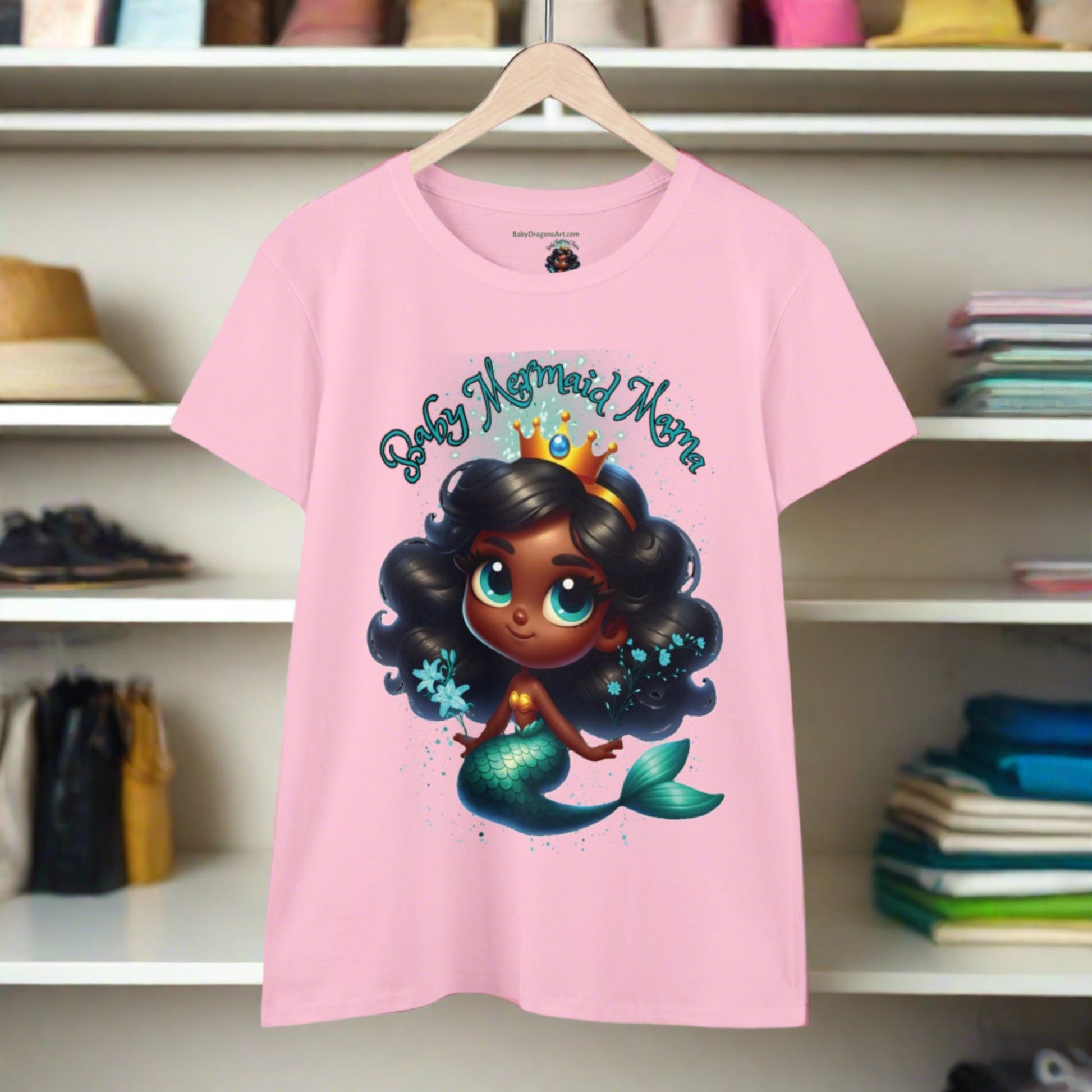 on a light pink tee a baby mermaid with black hair and a green tail wearing a gold crown