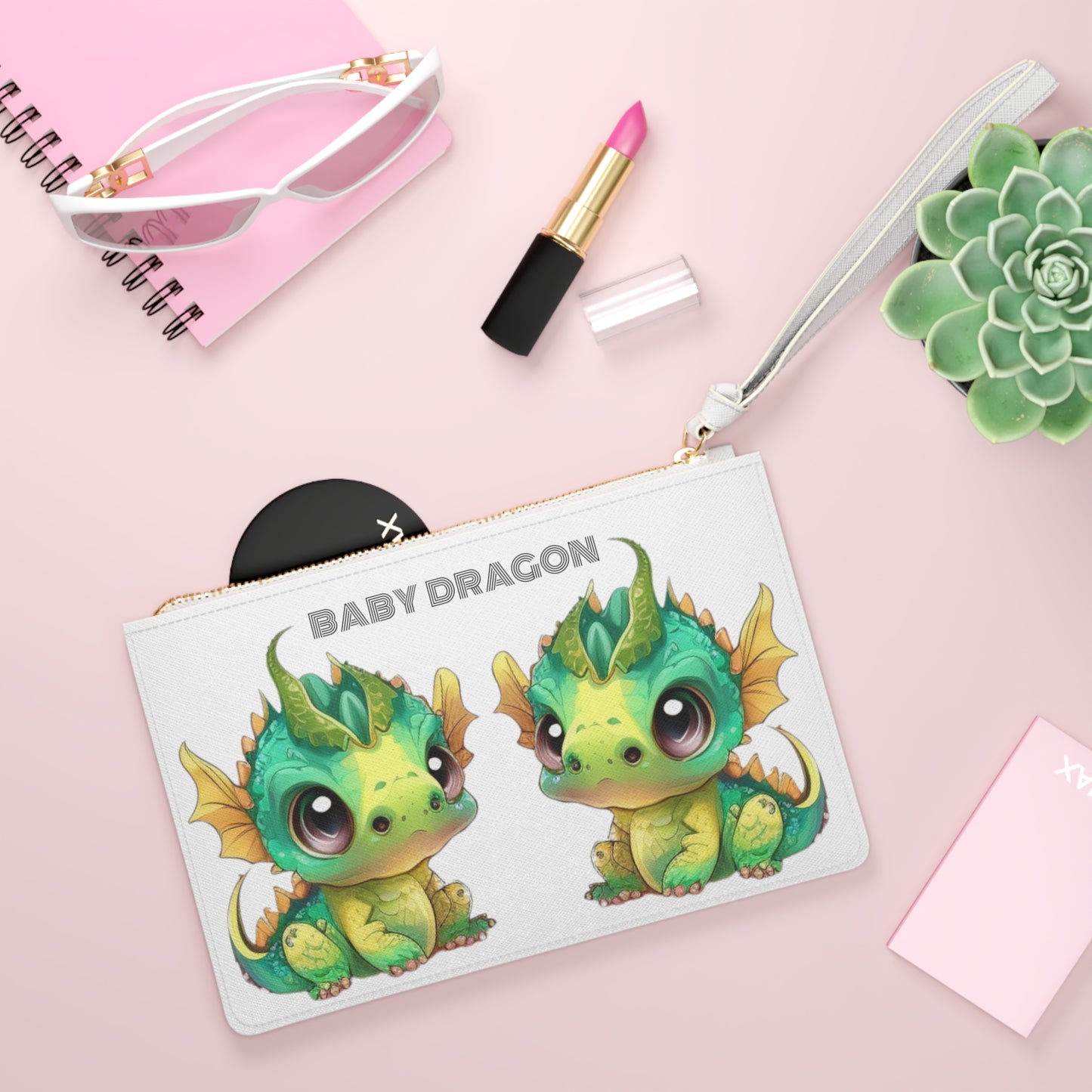 Baby Dragon is the decorative text above 2 darling baby Bobby dragons facing each other - Bobby is in greens and yellows with a little green horn popping out of his cute head - on a white vegan leather sofino patterned clutch bag with a loop white handle and gold zipper - excellent quality! On the other side of the clutch little Bobby dragons are all over it an inch apart.